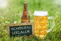 Urlaubsgrüße schönen Urlaub: Eine Tafel mit der Aufschrift 'Schönen Urlaub' steht auf einer Wiese neben einem Bierkrug und einer Flasche. Perfekt, um entspannte Urlaubsstimmung und kurze Urlaubsgrüße zu vermitteln.