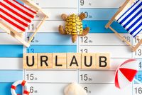 Kalender mit Abwesenheitsnotiz „Urlaub“ aus Holzbuchstaben und dekorativen Urlaub-Strandelementen wie Liegestühlen, Schildkröte und Sonnenschirm.