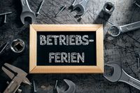 Tafel mit der Aufschrift „Betriebsferien“ umgeben von Werkzeugen auf einem Werkstatttisch eines Handwerkers.