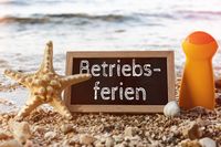 Tafel mit der Schrift Betriebsferien im Urlaub an einem Strand, umgeben von einem Seestern, einer Muschel und einer Sonnencremeflasche.