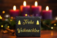 Schieferplatte mit Frohe Weihnachten Schriftzug vor brennenden Kerzen.