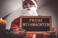 Der Weihnachtsmann hält ein Schild mit der Aufschrift 'Frohe Weihnachten' neben einer Kerze.