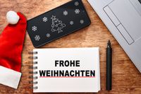 Ein Notizbuch mit Frohe Weihnachten Schriftzug neben einem Laptop, ideal für geschäftliche Weihnachtsgrüße.