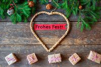 Frohes Fest wünscht ein geflochtenes Herz aus Stroh mit festlicher Tanne und kleinen Geschenken auf einem rustikalen Holztisch.