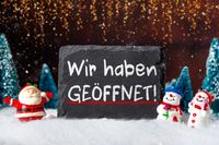 Schiefertafel mit 'Wir haben geöffnet!' umgeben von Weihnachtsfiguren im Schnee.