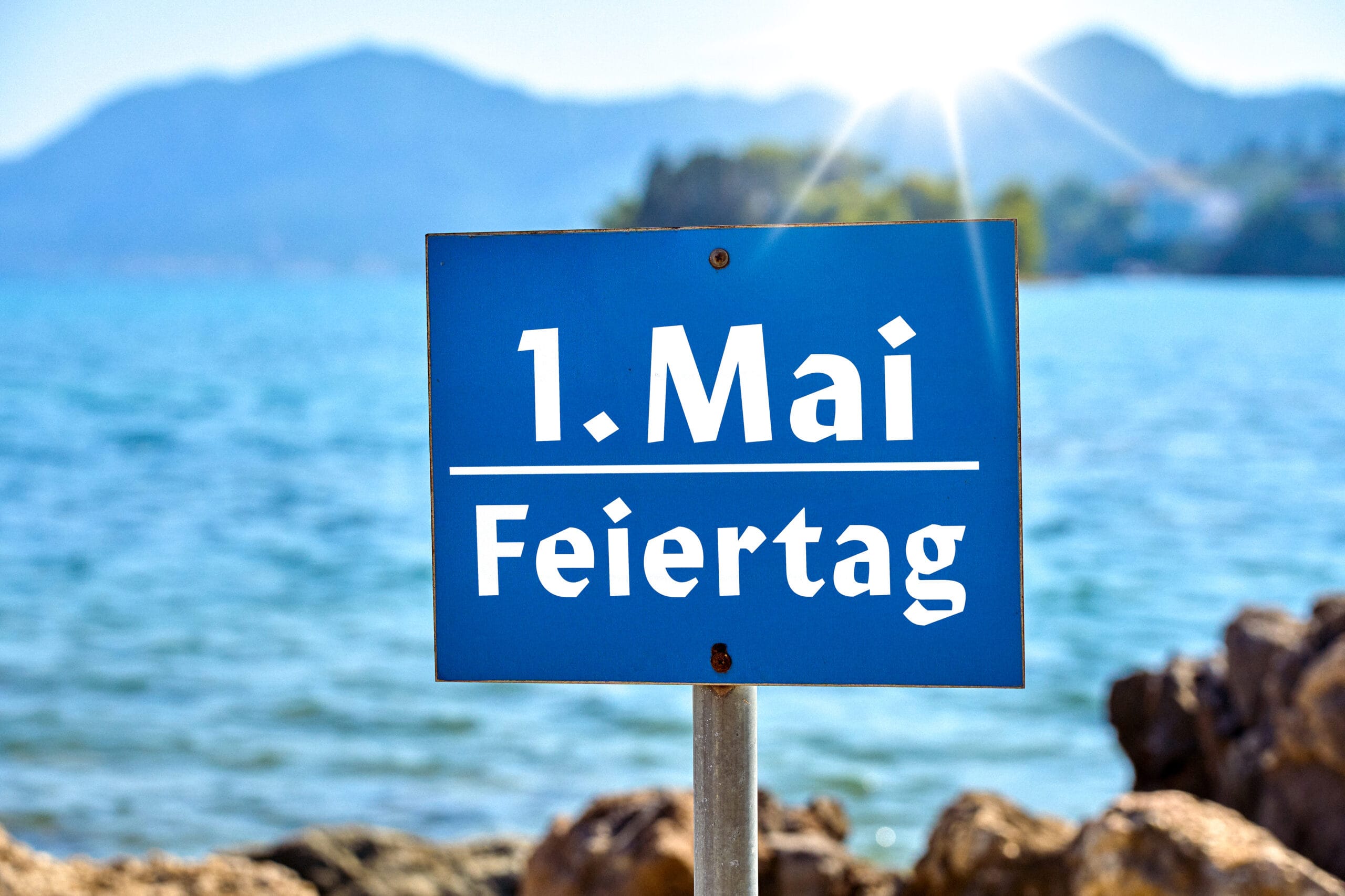 Blaues hinweisschild mit aufschrift 1. Mai feiertag vor einer küstenlandschaft mit meer und bergen
